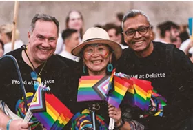 Three people smiling towards camera with pride flags in foreground