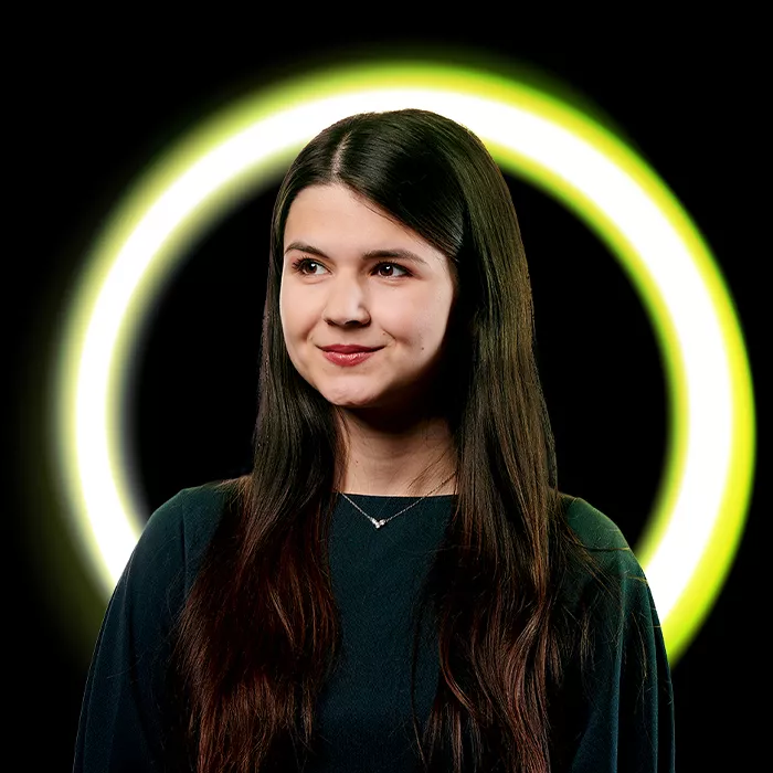 Mladá žena s dlouhými hnědými vlasy stojí před černo-zeleným kruhovým světelným pozadím