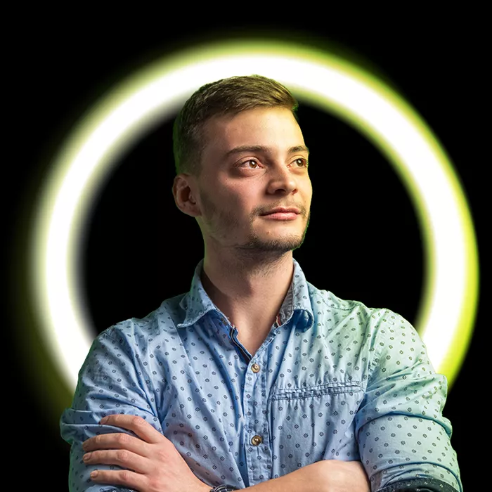 Mladý muž ve světle modré košili s překříženýma rukama stojí před černo-zeleným kruhovým světelným pozadím