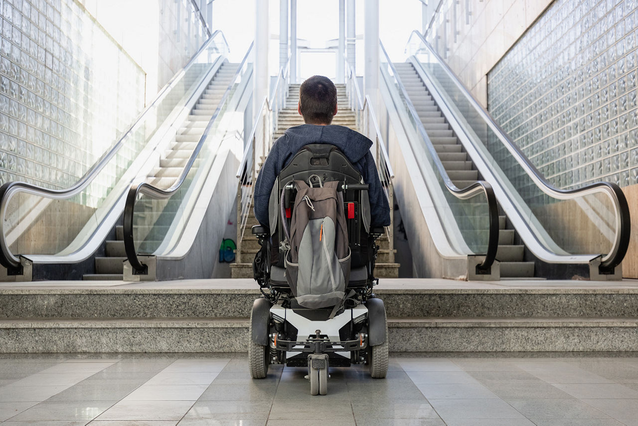A person using a motorized wheelchair at the bottom of stairs and escalators looking up at the street above.